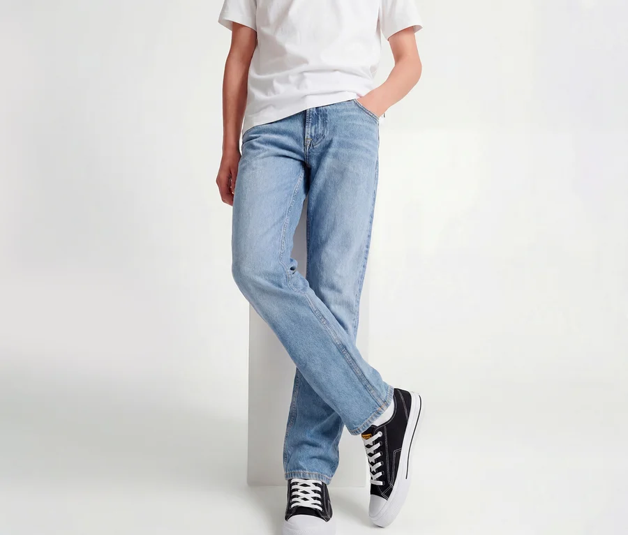 Person trägt ein weißes T-Shirt, hellblaue Jack&Jones JJOriginal Chris Jeans und schwarze Sneaker, lehnt mit gekreuzten Beinen an einem weißen Block.