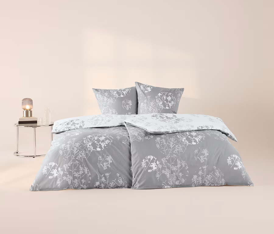 Graue Renforcé-Bettwäsche mit weißem Blumenmuster, bestehend aus zwei Kissen und zwei Decken, wobei eine zurückgeschlagen ist. Links steht ein runder Glastisch mit einer Lampe und einer Flasche.