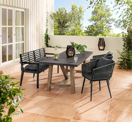 Eine Gartensitzgruppe mit einer Gartenbank mit Armlehne und einer Sun Garden Gartenbank-Auflage, einem Esstisch mit Polyzement-Tischplatte und Dining-Sesseln mit Textilgeflecht. Auf dem Tisch steht eine Solar-Laterne mit Textilband.