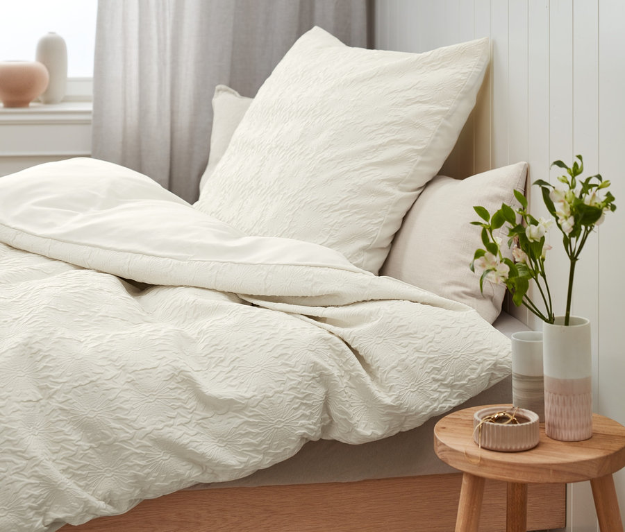 Nahaufnahme von beiger Struktur-Bettwäsche (Normalgröße, Doppelgröße, Übergröße) auf einem Bett mit Holzrahmen. Auf einem Nachttisch steht eine Vase mit Blumen.