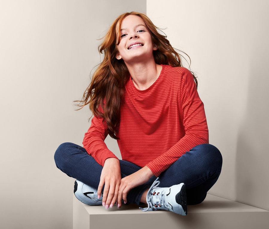 Mädchen sitzt mit gekreuzten Beinen auf einem Sockel und trägt ein Langarmshirt.