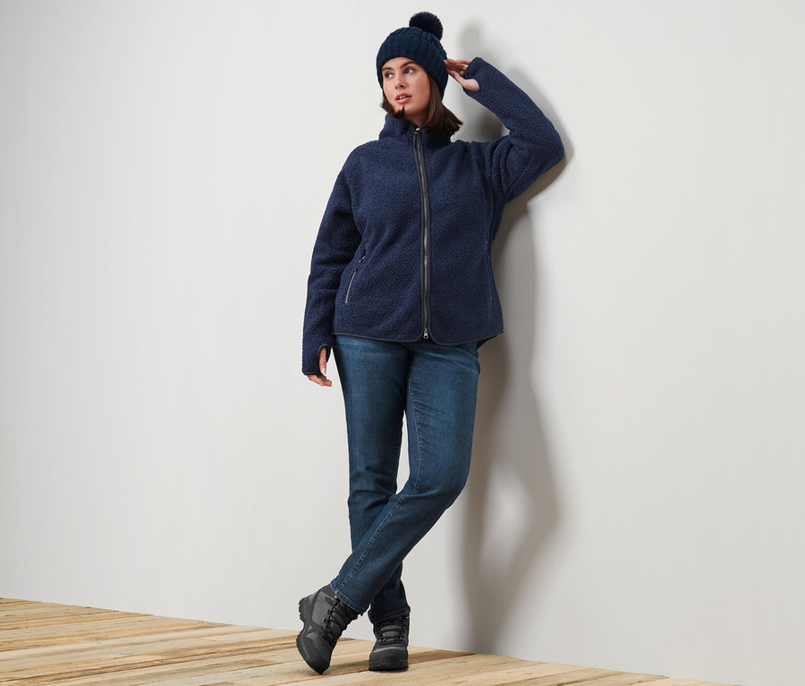 Frau posiert an einer Wand, trägt eine blaue Teddy-Jacke, eine Slim-Denim »Fit Emma«, darkblue Denim, und eine Strickmütze.