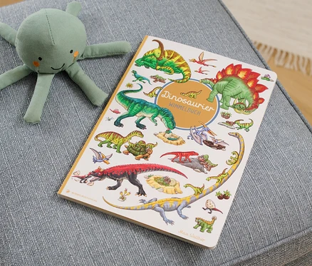 Auf einem grauen Hocker liegen ein »Dinosaurier Wimmelbuch« und ein grünes Tintenfisch Stofftier.