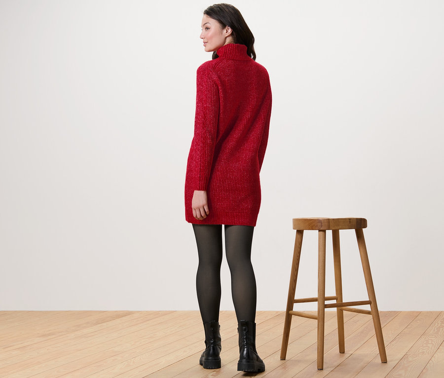 Frau trägt langen Strickpullover mit Zopfmuster in Rot, schwarze Strumpfhose und Stiefel, steht neben einem Holzstuhl.