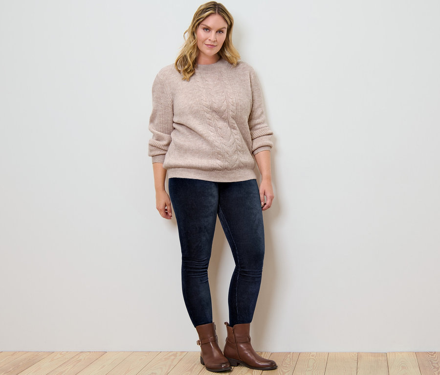 Frau posiert in Grobstrickpullover mit Zopfmuster und Cord-Leggings.