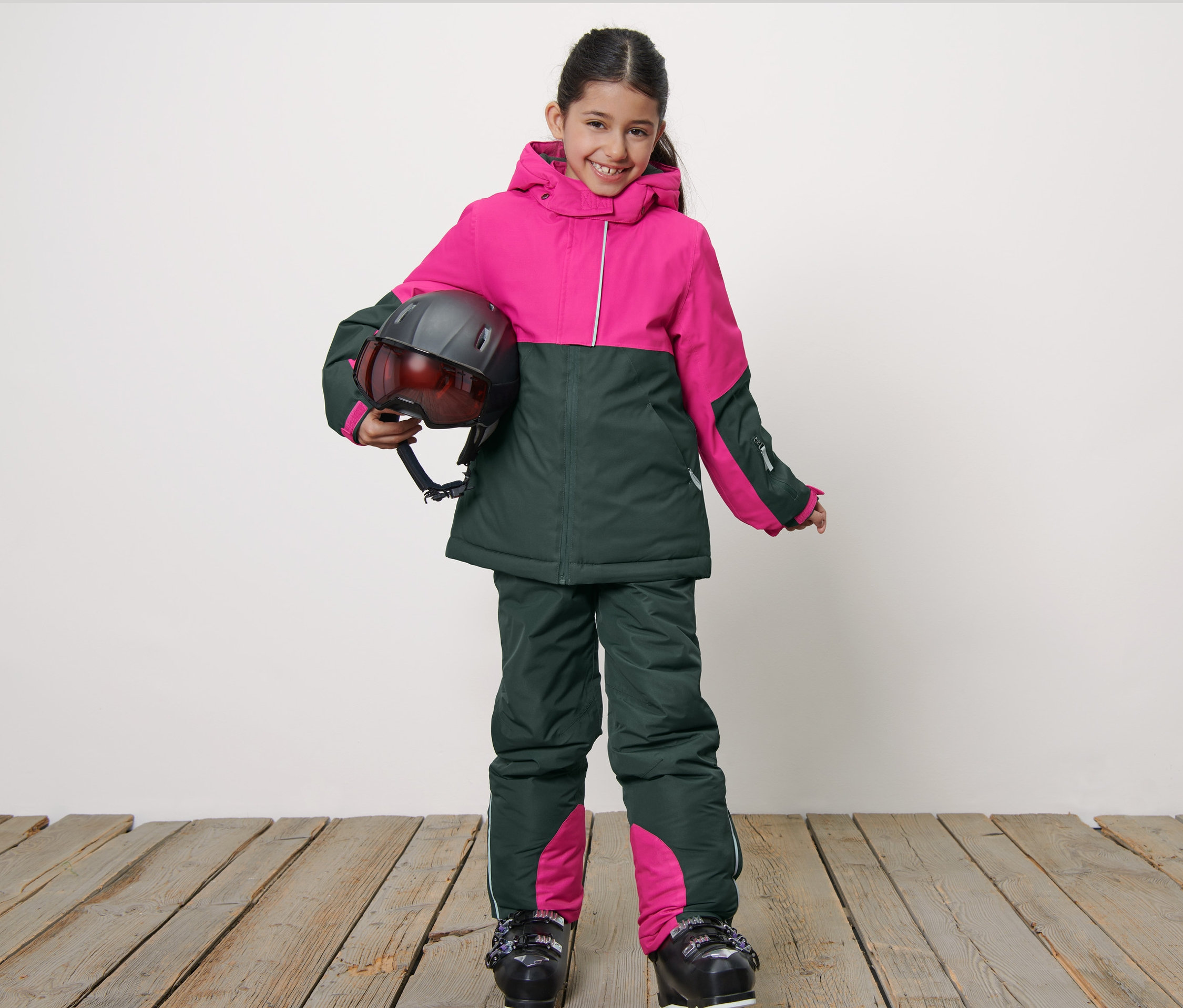 Ein lächelndes Mädchen in einer Kinder-Skijacke hält einen Helm.