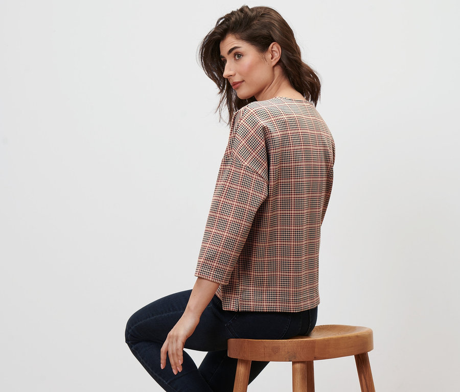 Frau sitzt auf einem Holzhocker und trägt ein Sweatshirt mit 3/4-Arm im Hahnentrittmuster.