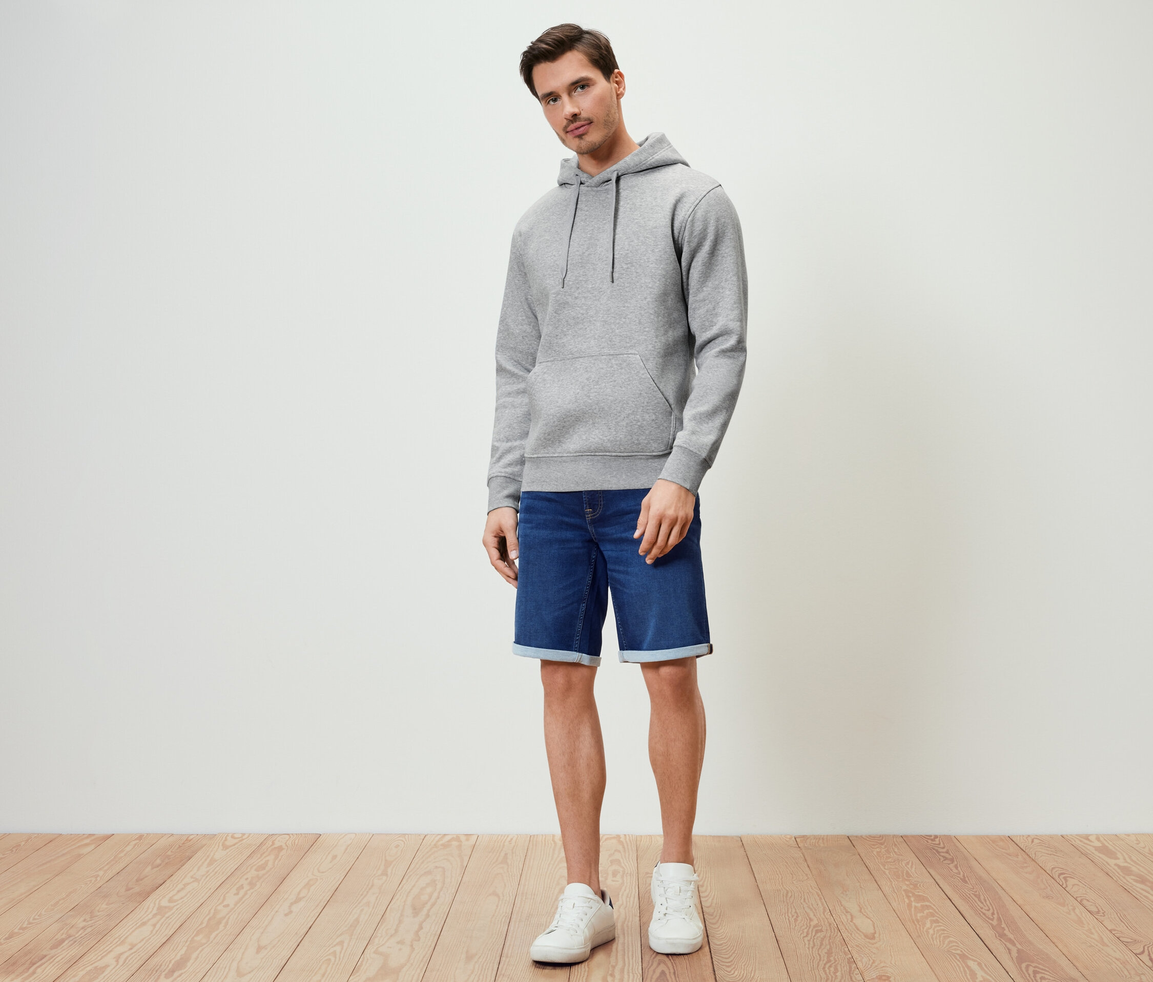 Mann steht und trägt einen grau melierten Hoodie, Jeansshorts und weiße Turnschuhe.