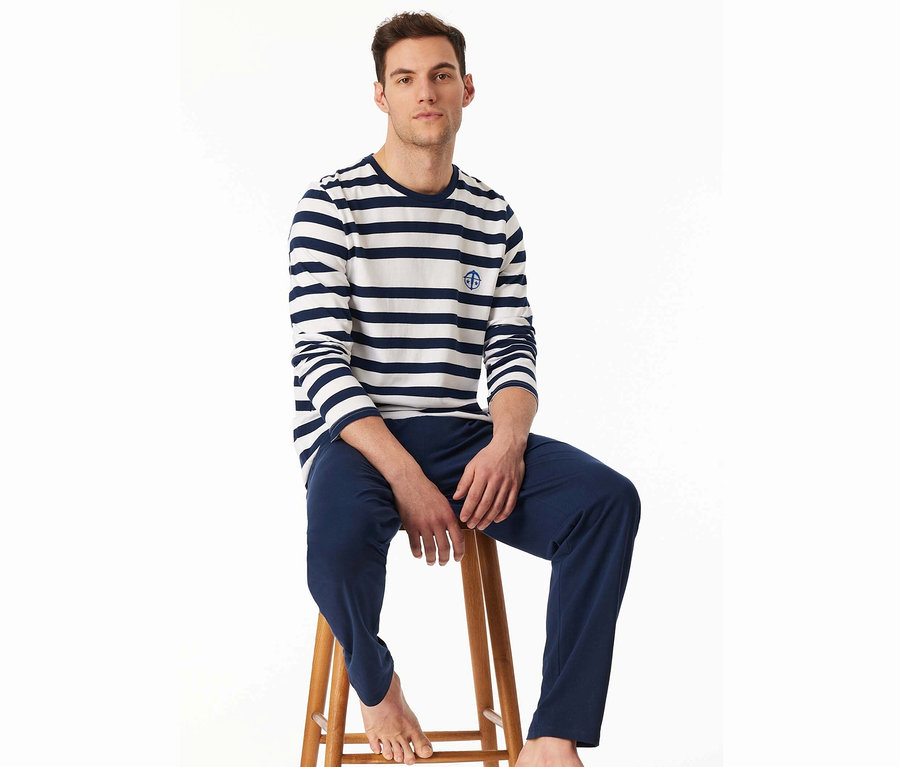 Mann sitzt auf einem Holzhocker, trägt ein gestreiftes Langarmshirt und blaue Hosen.