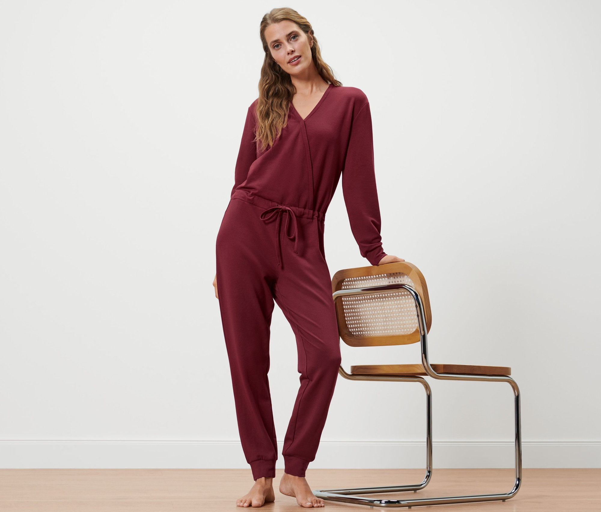 Frau posiert in weinrotem Loungeoverall, eine Hand auf einem Stuhl.