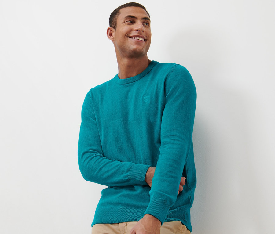 Ein lächelnder Mann trägt einen aqua Pullover mit Rundhalsausschnitt.