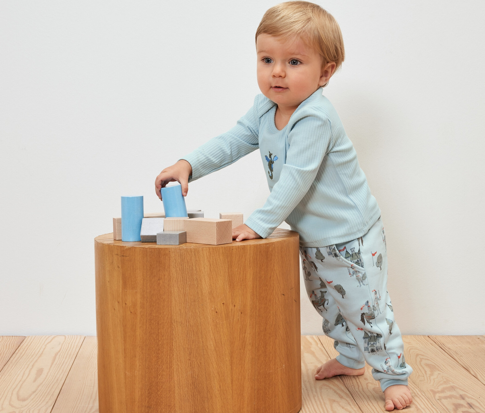 Baby spielt mit Holzbausteinen auf einem Holztisch, trägt ein Outfit mit Rittermotiv und eine 2 Baby-Sweathosen, Ritter.