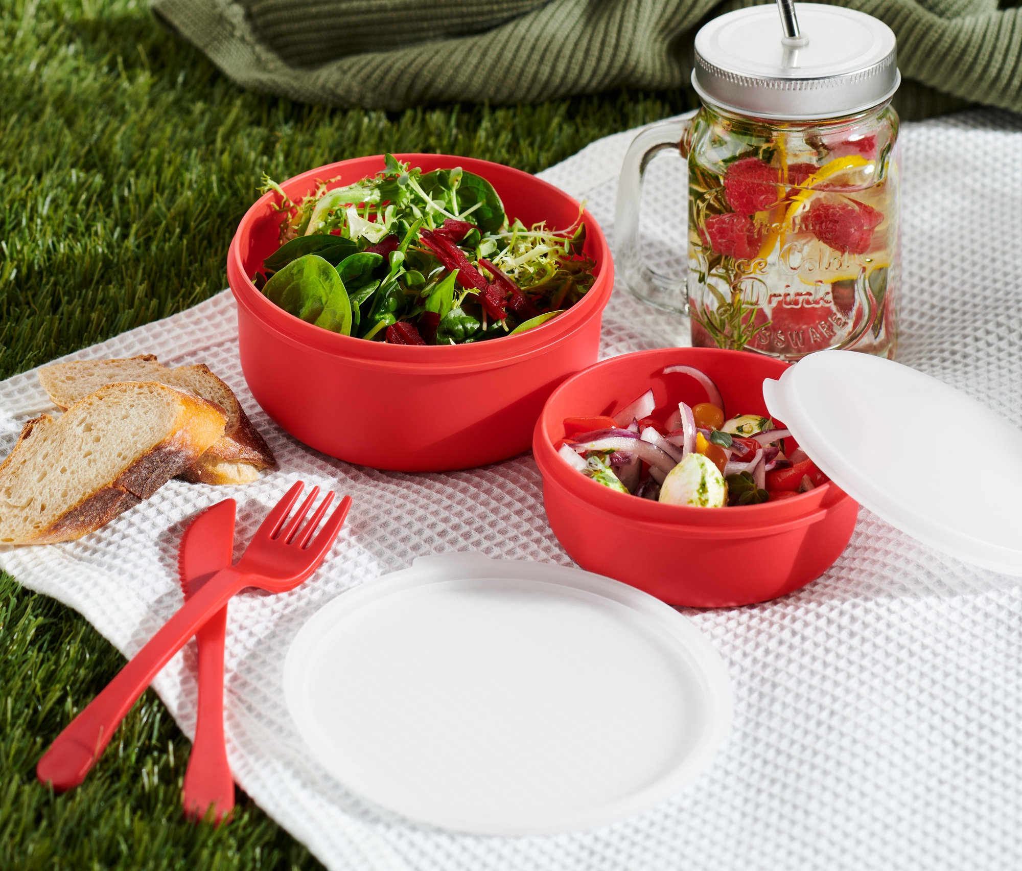 Auf einer karierten Decke liegen zwei rote Schüsseln mit Deckel, Brot, Besteck und ein Glas mit Früchten.