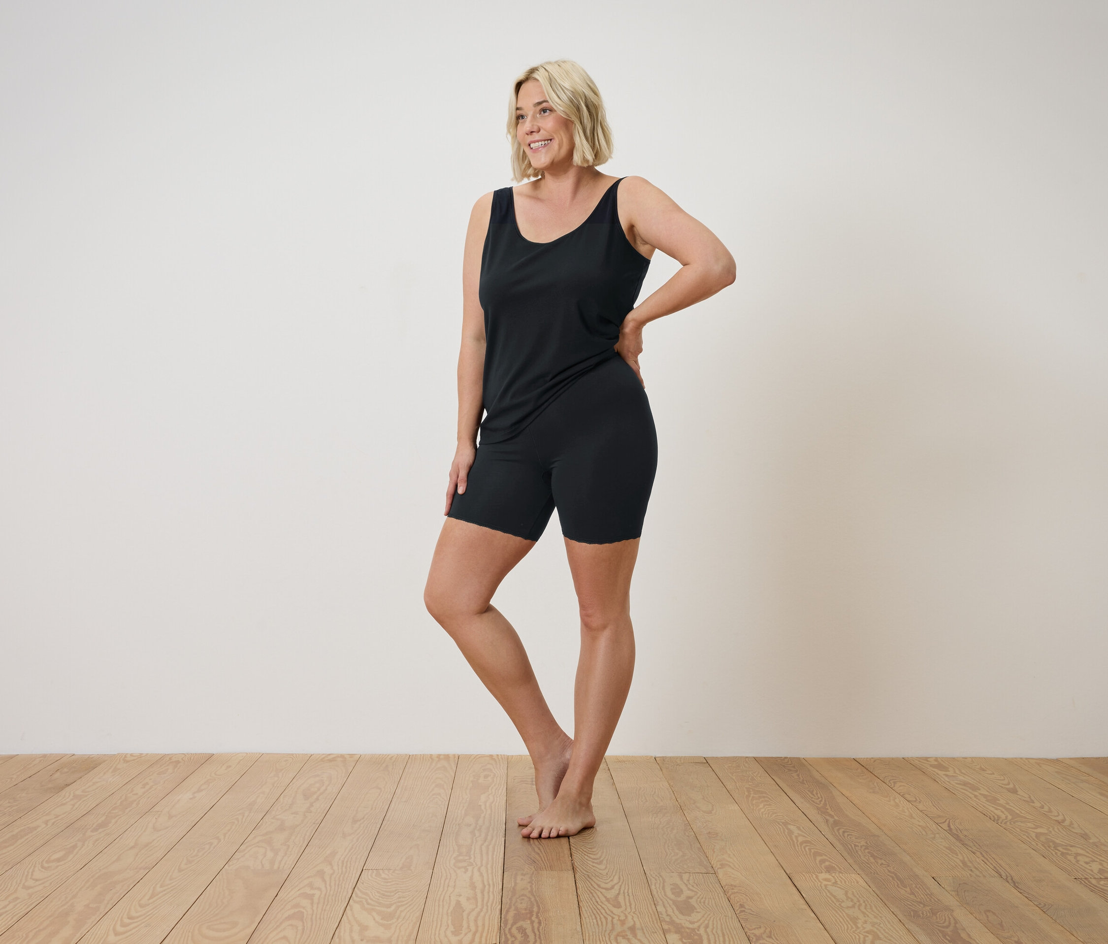 Blonde Frau in schwarzem Tanktop und Shorts posiert auf einem Holzboden.