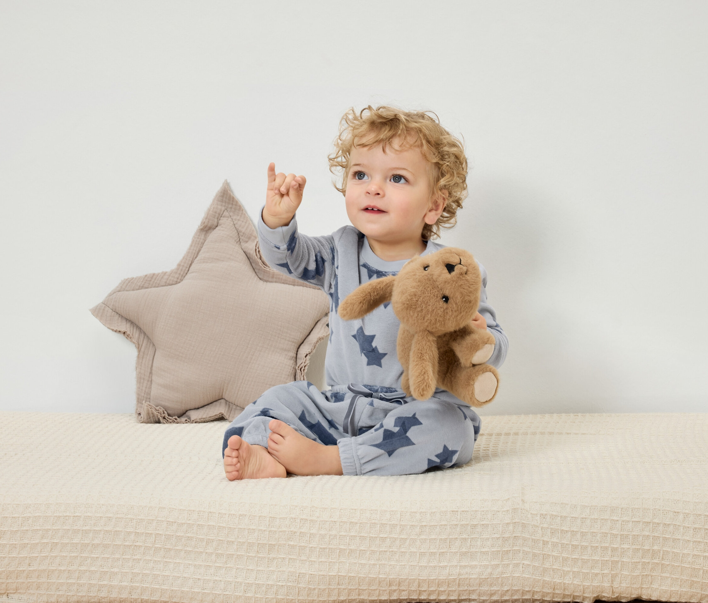 Ein Kind sitzt auf einer beigen Unterlage, hält ein Plüschkaninchen und zeigt mit dem Finger nach oben. Hinter ihm liegt ein sternförmiges Kissen.