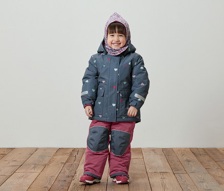 Ein lächelndes kleines Mädchen steht auf einem Holzboden und trägt eine blaue Winterjacke mit Herzmuster, eine rosafarbene Hose und ein Kopftuch.