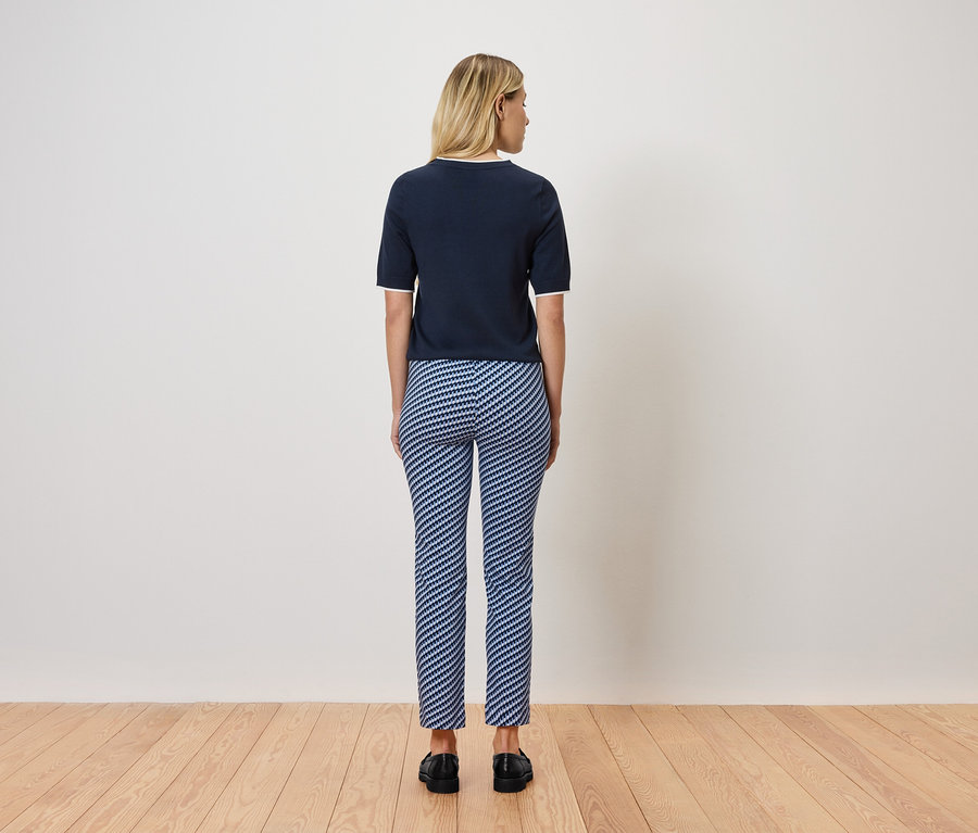 Blonde Frau steht mit dem Rücken zur Kamera, trägt ein marineblaues Kurzarmtop und blau-weiß gestreifte Hosen. Sie steht auf einem Holzboden.