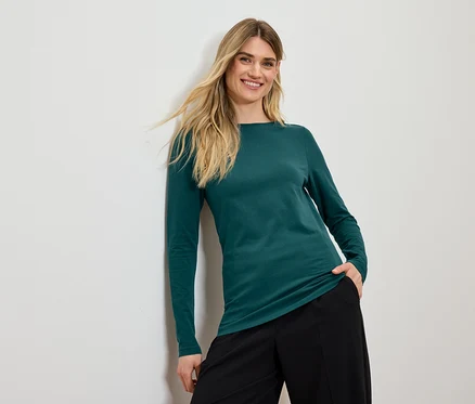 Lächelnde Frau mit blonden Haaren posiert in einem grünen Langarmshirt und einer schwarzen Hose.