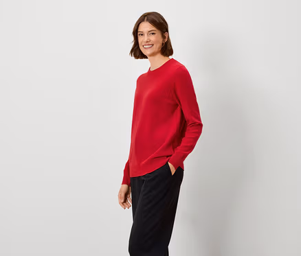 Frau mit braunen Haaren steht und trägt einen roten Pullover und eine schwarze Hose.
