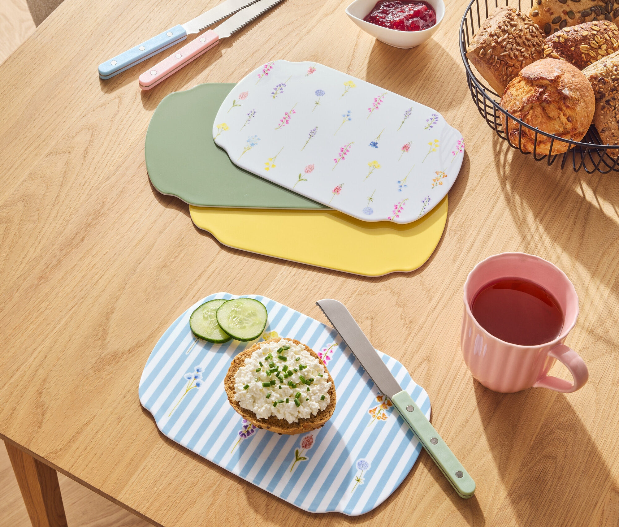 Auf einem Holztisch liegen Brote in einem Korb, Marmelade, Messer und Schneidebretter in verschiedenen Farben.