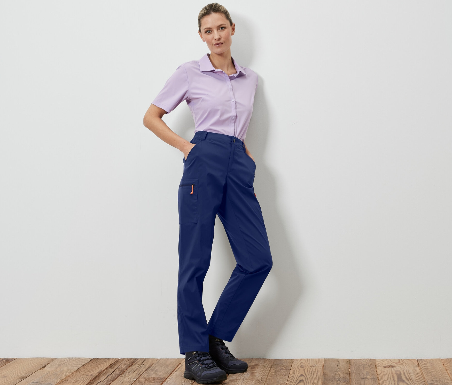 Eine Frau posiert in dunkelblauen, packbaren Outdoorhosen und einem helllila Hemd.