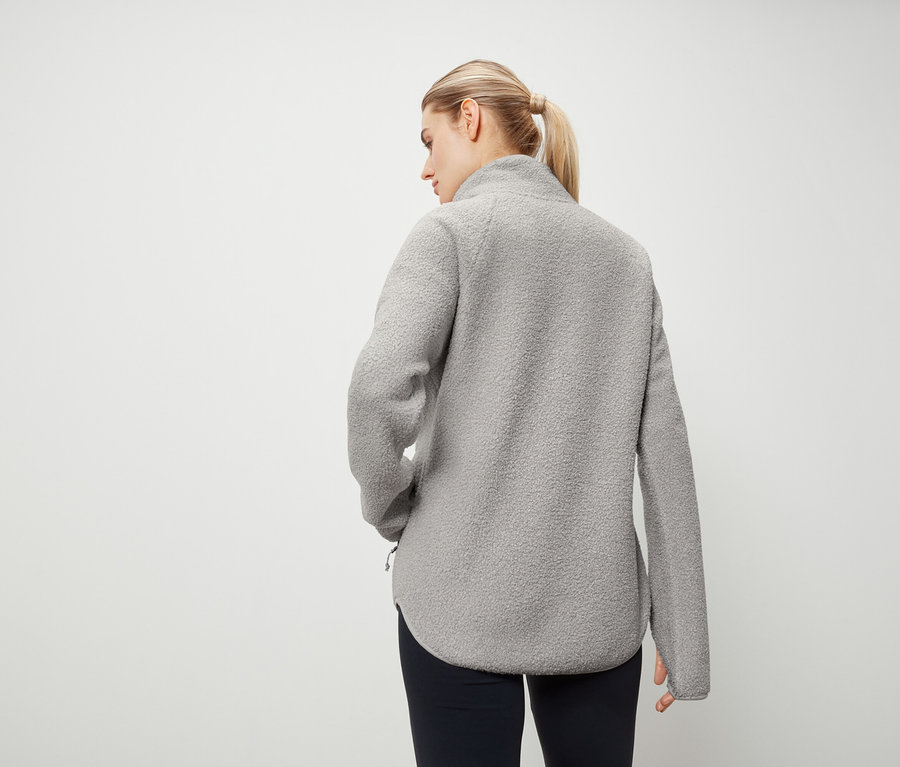 Von hinten trägt eine Frau mit blonden, zu einem Pferdeschwanz zusammengebundenen Haaren eine graue Fleecejacke und marineblaue Hosen.