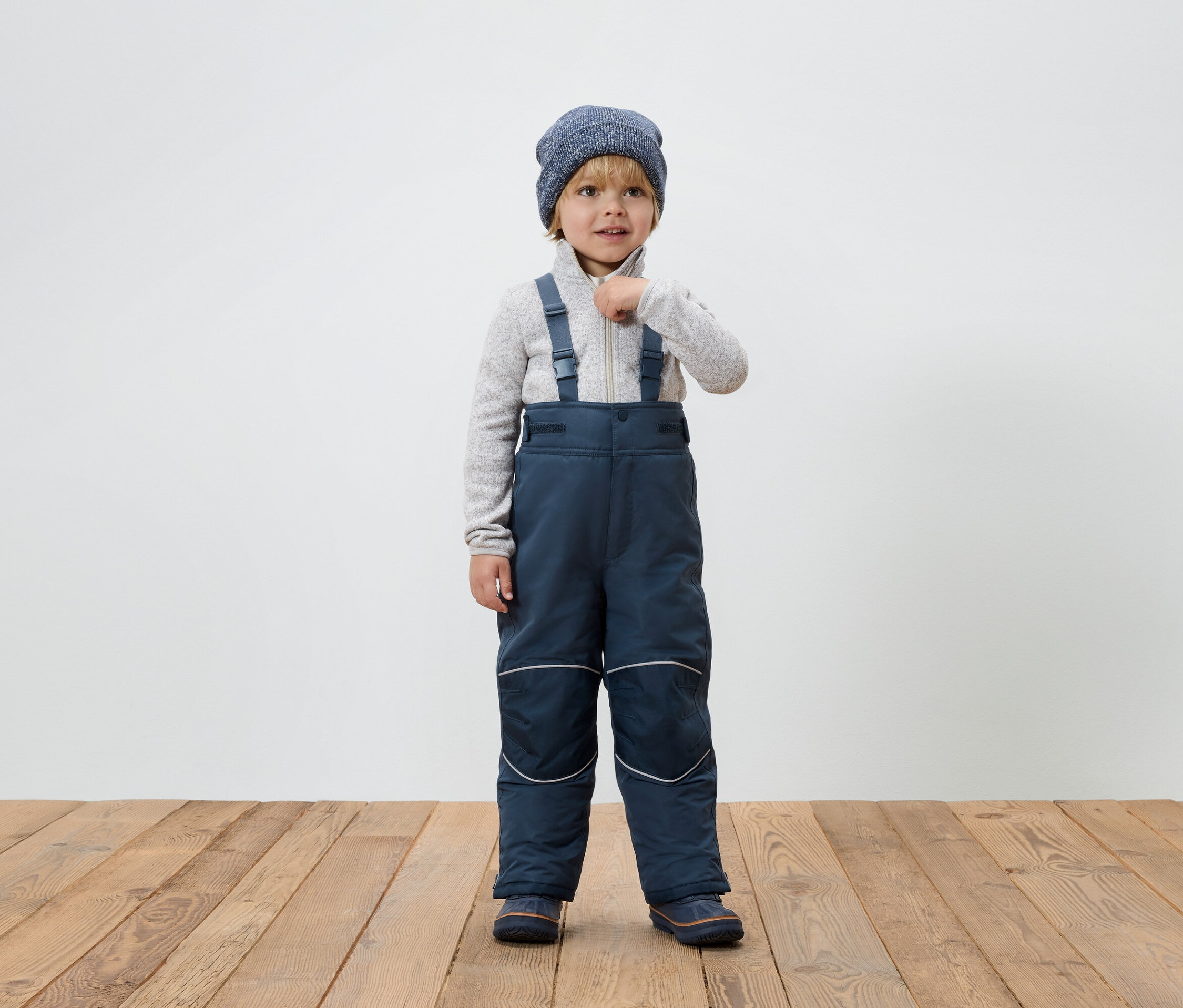 Ein Junge in blauer Winterkombination mit Hosenträgern und Strickmütze steht auf einem Holzboden und schließt seine Strickjacke.