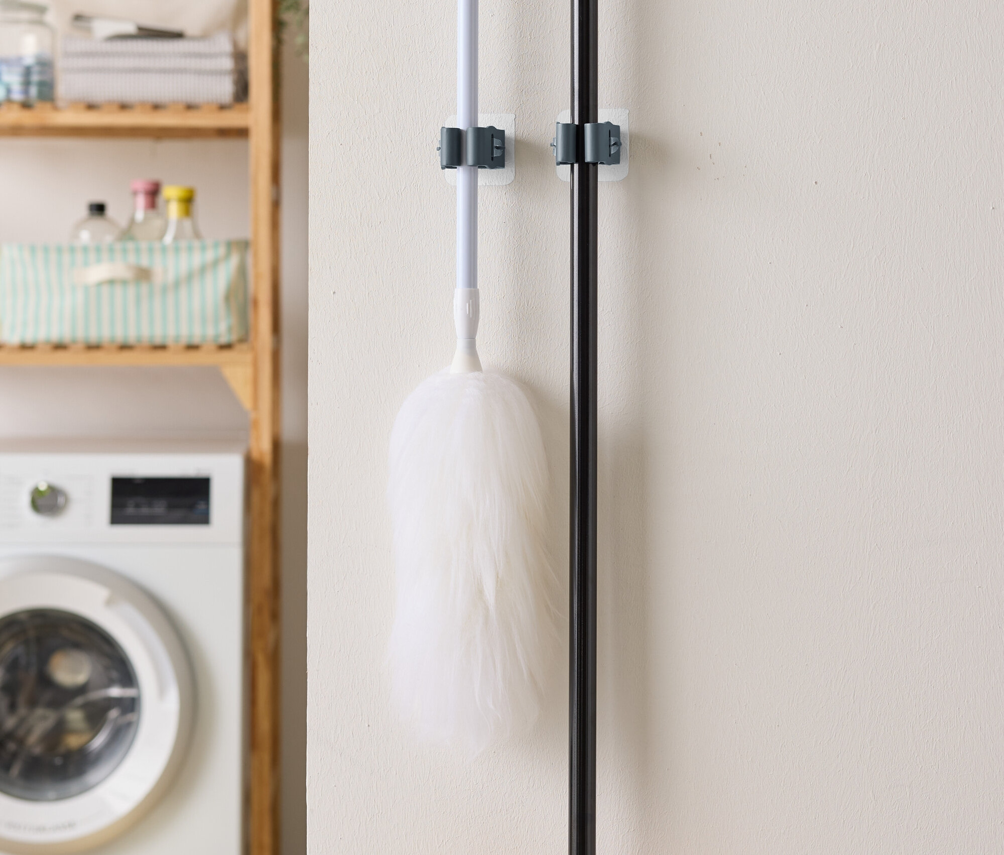 Ein Staubwedel und ein Besen hängen an der Wand, im Hintergrund eine Waschmaschine und ein Regal.