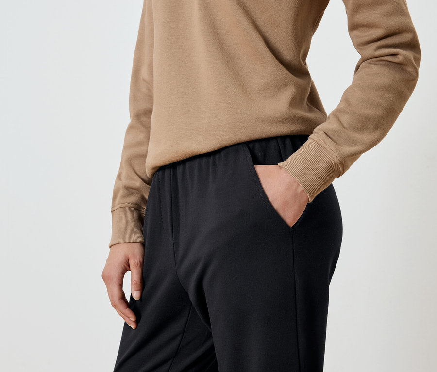 Detailaufnahme einer Frau mit der Hand in der Tasche einer schwarzen Jogginghose und einem beigen Sweatshirt.