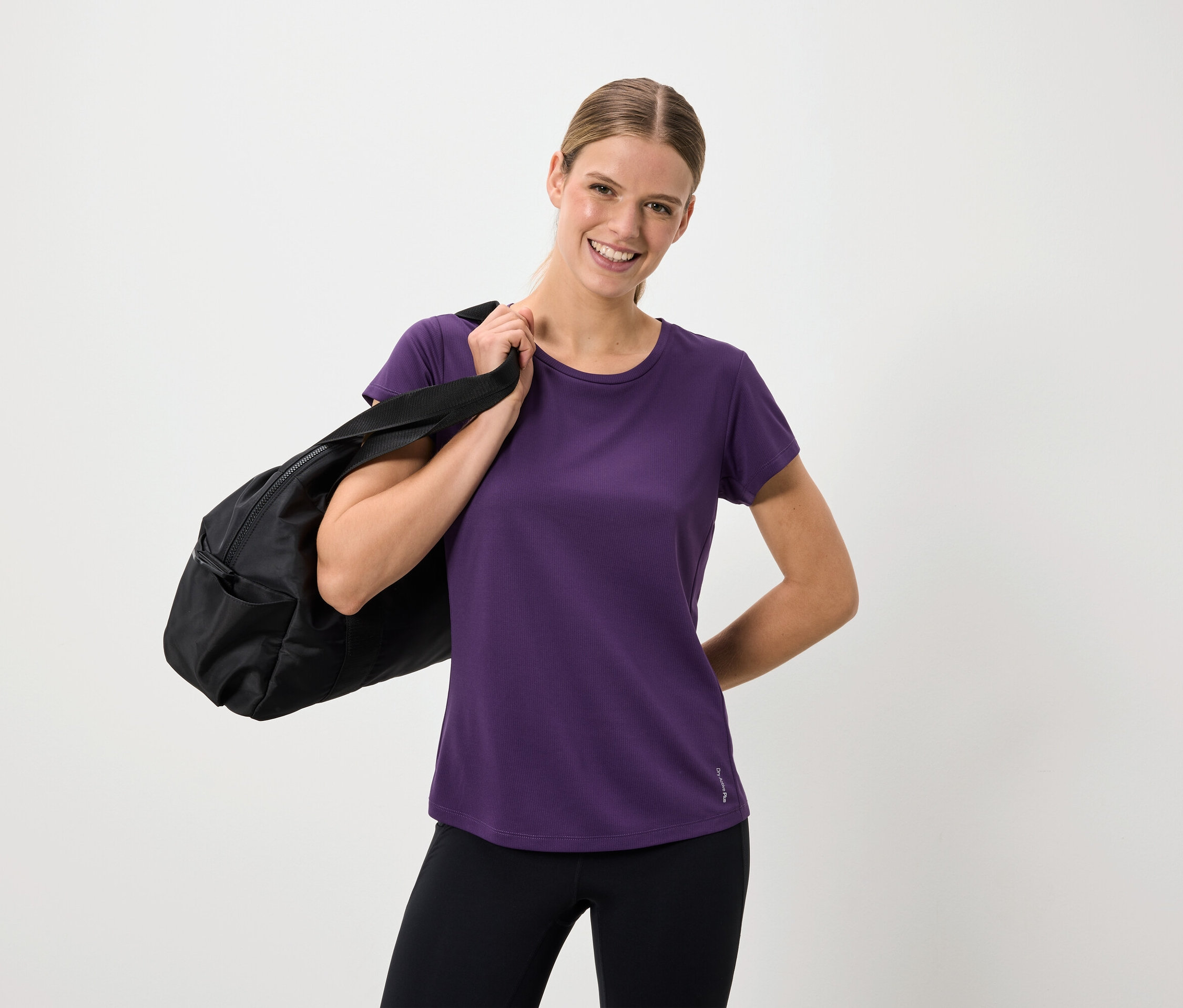 Lächelnde Frau in lila T-Shirt und schwarzen Leggings mit einer schwarzen Tasche über der Schulter.