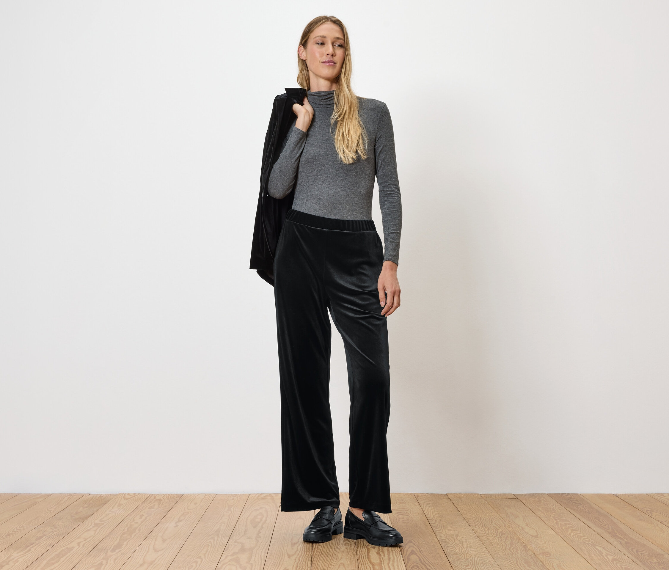 Frau posiert in grauem Langarmshirt und schwarzer Samthose, Jacke über der Schulter.