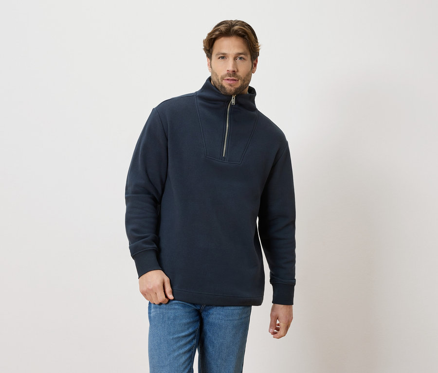 Ein Mann trägt einen marineblauen Pullover mit Reißverschluss und blaue Jeans.