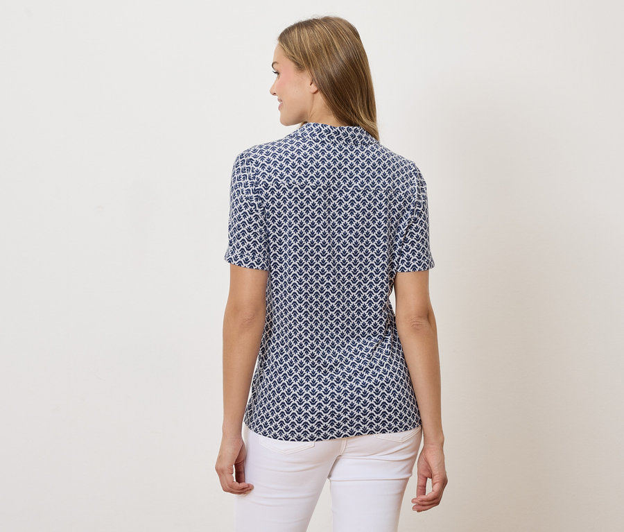 Rückansicht einer Frau mit blonden Haaren, die eine blau-weiß gemusterte kurzärmlige Bluse und weiße Jeans trägt.