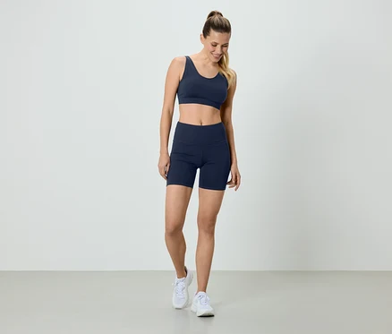 Frau in marineblauem Sport-BH und Radlerhose, mit weißen Turnschuhen, stehend vor einem weißen Hintergrund.
