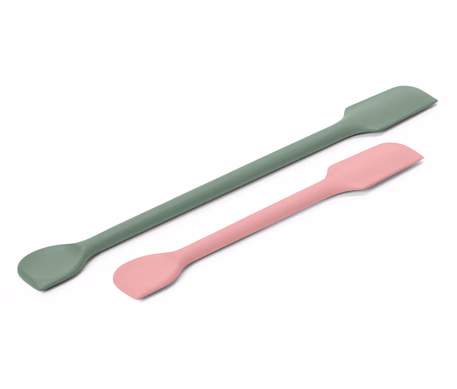 Ein grüner und ein rosa Spatel liegen diagonal auf einem weißen Hintergrund.