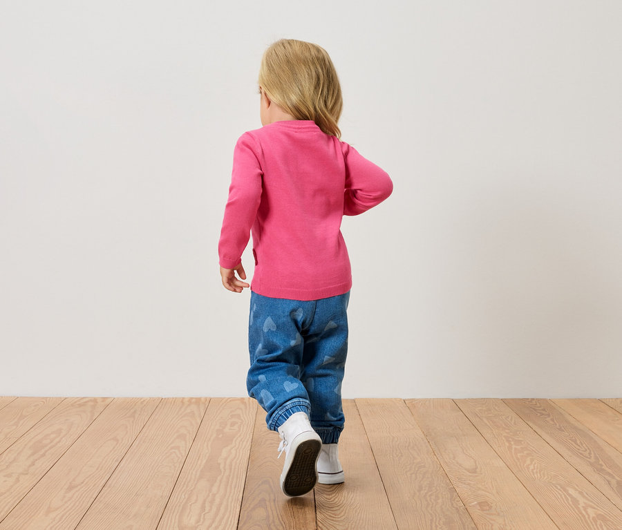 Kleines blondes Kind von hinten, trägt einen pinken Pullover, blaue Jeans mit Herzen und weiße Schuhe, geht auf einem Holzboden.