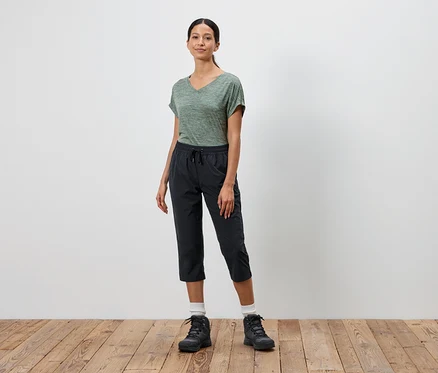 Frau steht vor einer weißen Wand und trägt ein grünes T-Shirt, schwarze Dreiviertelhosen und schwarze Wanderschuhe.