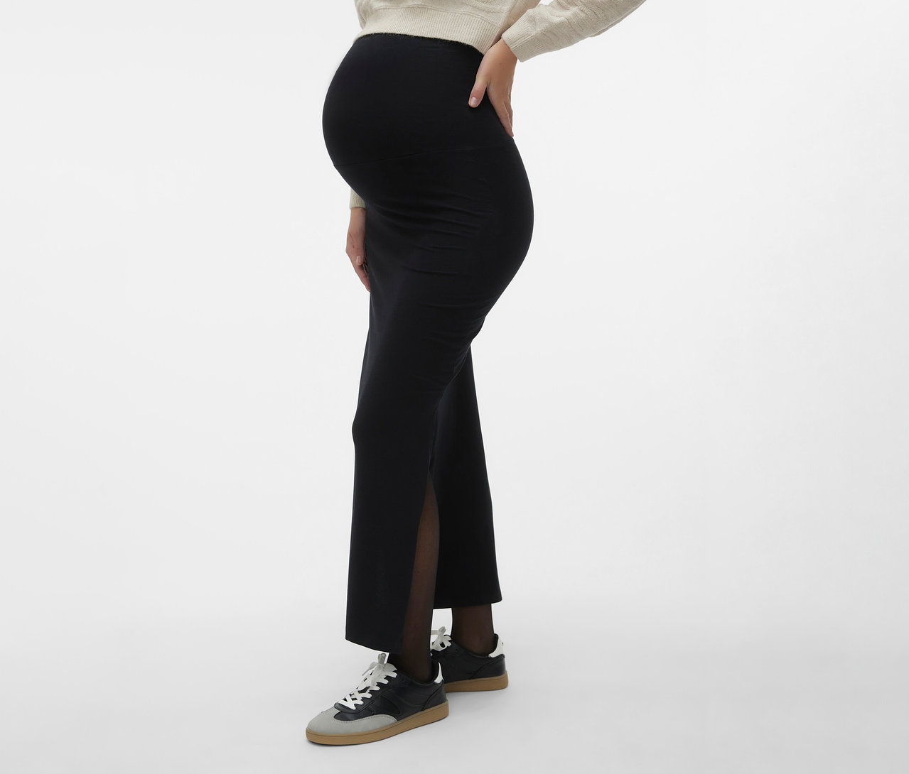 Schwangere trägt MAMALICIOUS Maxirock »MLEMMA TUBE« mit Seitenschlitz, dunkle Strumpfhose und Turnschuhe vor neutralem Hintergrund.