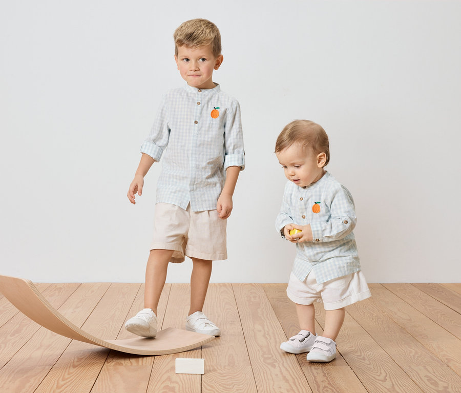 Zwei Kinder, ein älterer Junge, der auf einem Holzwippe steht, und ein kleiner Junge, der einen gelben Ball hält, beide tragen karierte Hemden mit Orangenapplikation.