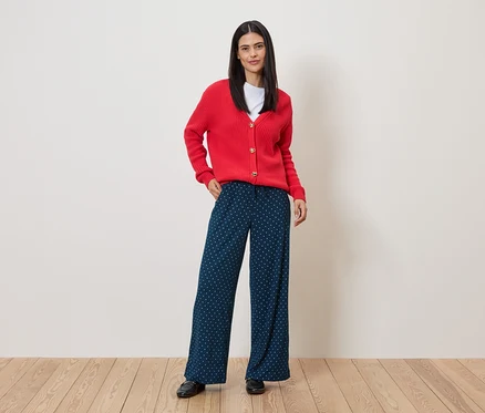 Frau mit roter Strickjacke und blau-weiß gepunkteter Hose steht auf Holzboden vor einer Wand.