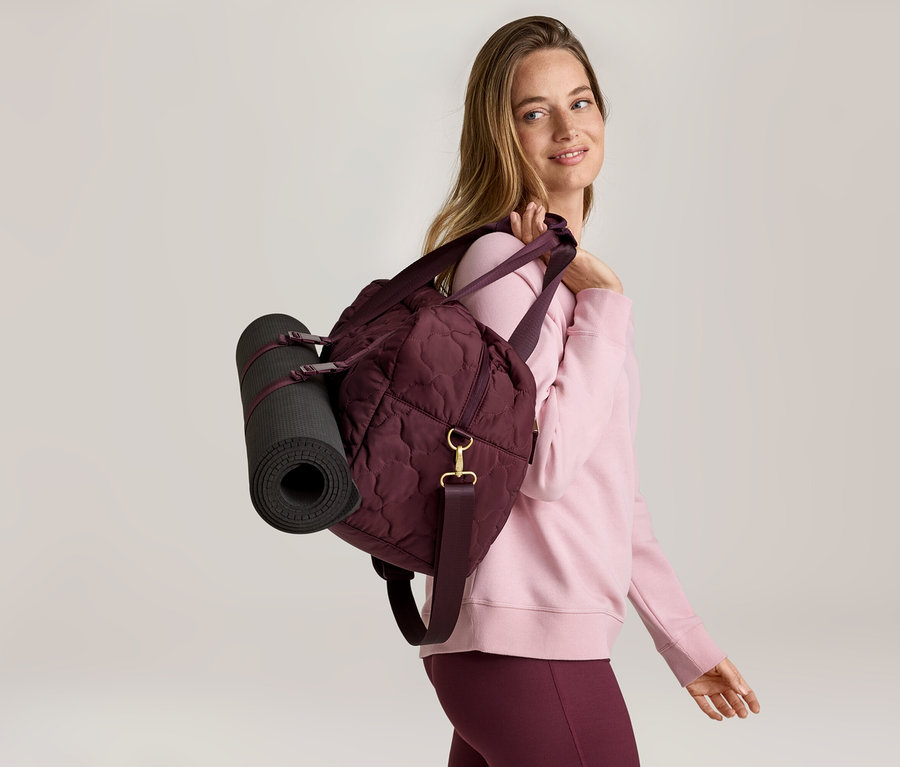 Frau mit Sporttasche und Yogamatte über der Schulter, trägt rosa Pullover und bordeauxfarbene Leggings.