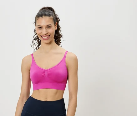 Frau mit hochgesteckten lockigen Haaren, die einen pinkfarbenen Sport-BH und marineblaue Leggings vor einem weißen Hintergrund trägt.