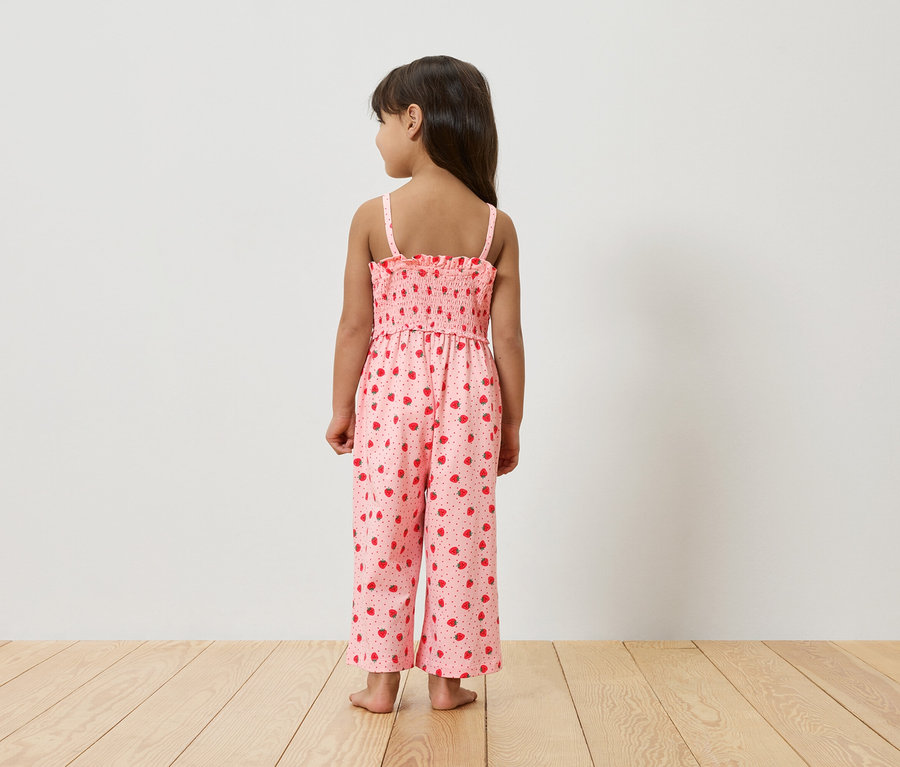 Ein junges Mädchen steht mit dem Rücken zur Kamera und trägt einen rosa Jumpsuit mit Erdbeermuster.
