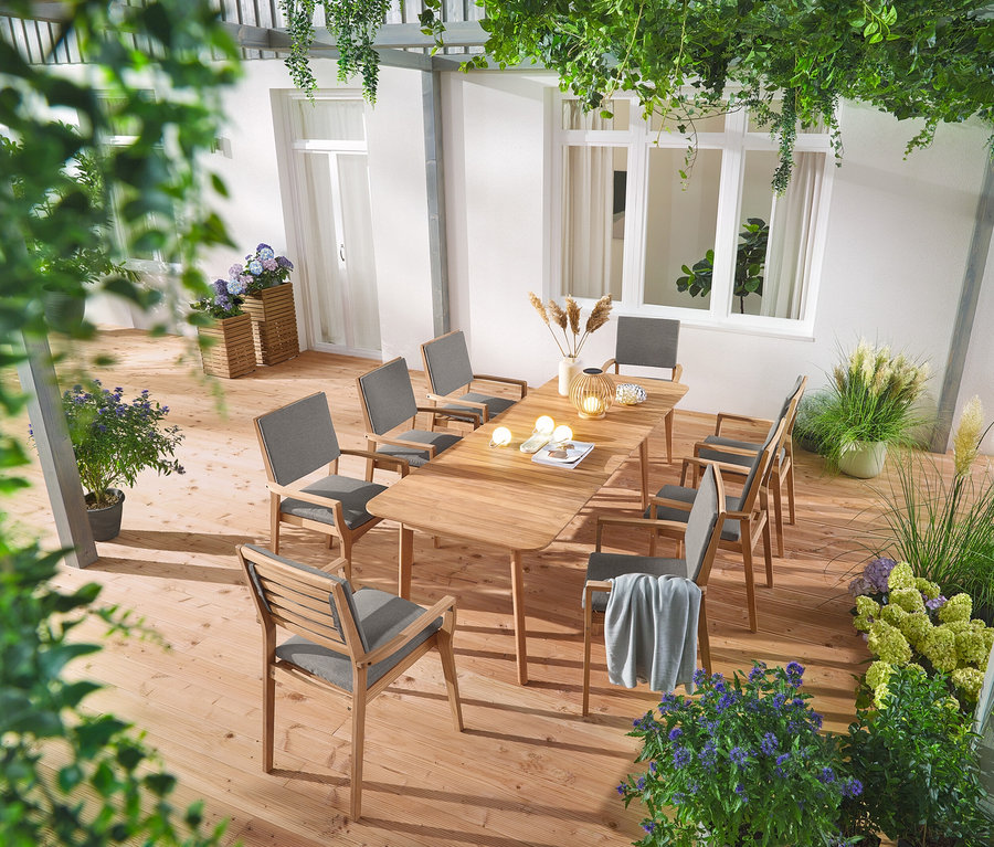 Ein ausziehbarer Teak-Esstisch mit acht stapelbaren Teak-Gartenstühlen steht auf einer Terrasse, dekoriert mit zwei Pflanzsäulen »Tinus«, einer runden Solar-Laterne aus Holz und drei Solar-Spießen »Hortensie«.
