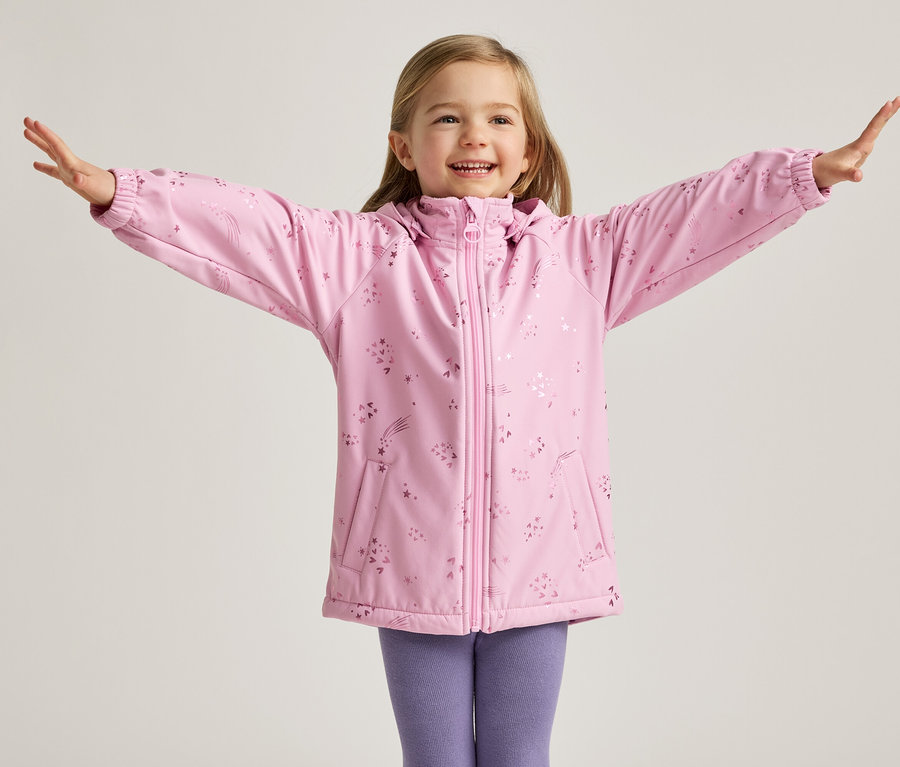 Ein lächelndes Mädchen mit blonden Haaren steht mit ausgestreckten Armen in einer rosa Jacke mit Sternen und lila Leggings vor einem hellen Hintergrund.