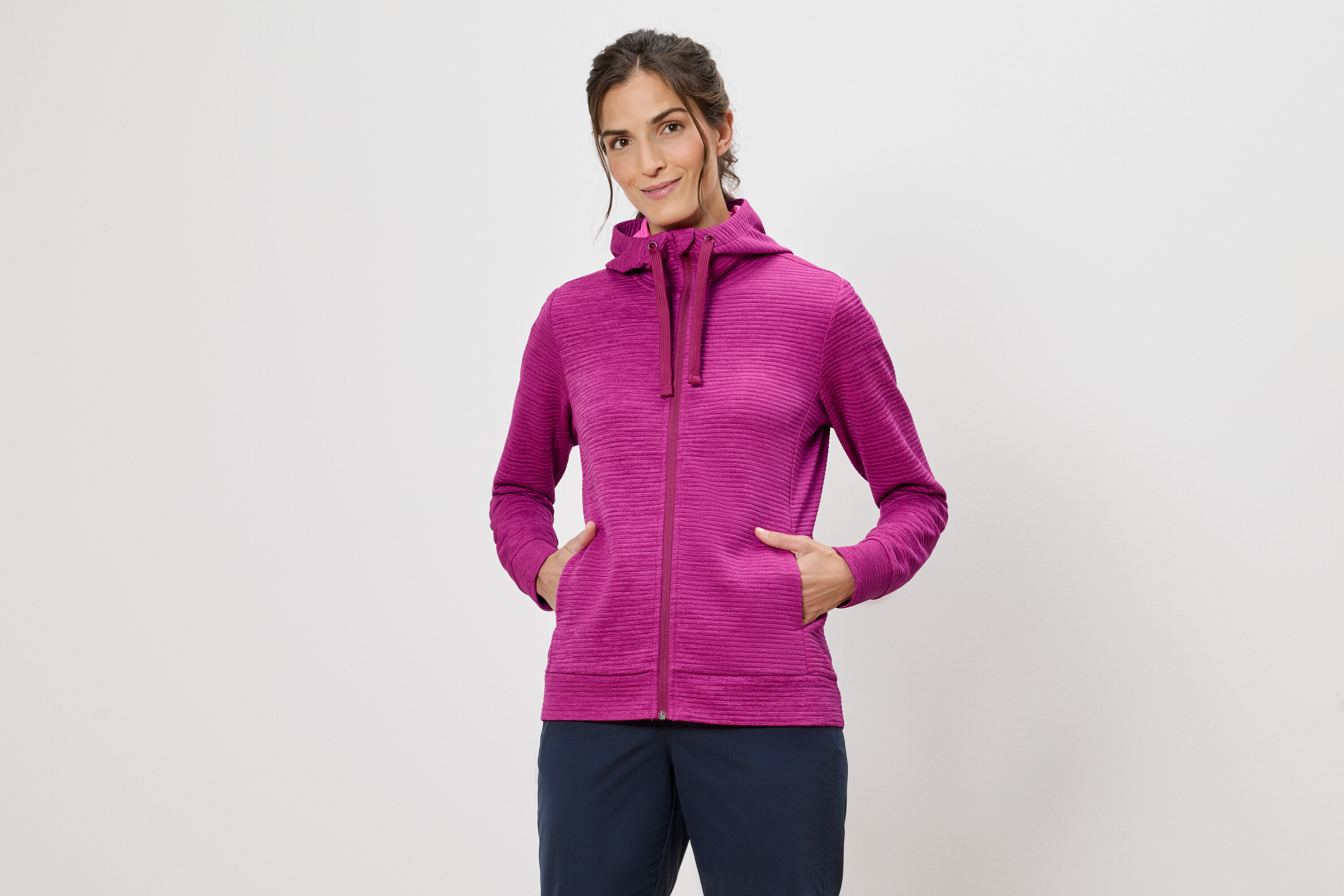 Frau mit Kapuzenpullover in Pink mit Händen in den Taschen vor einem weißen Hintergrund.