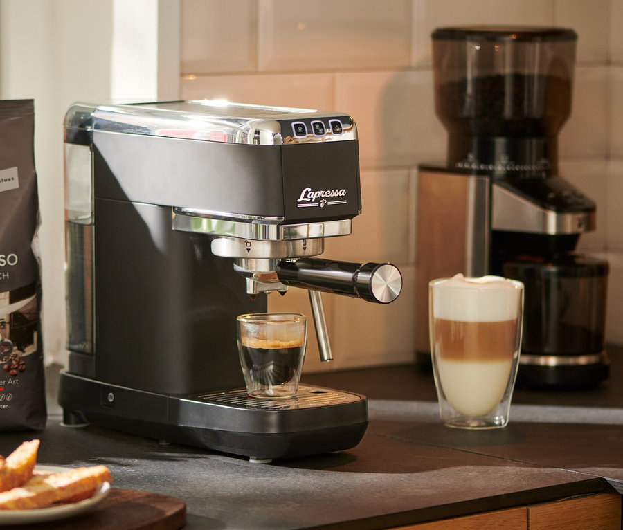 Espresso läuft aus einer »Lapressa« Siebträgermaschine, schwarz, in ein Glas. Im Hintergrund stehen eine Kaffeemühle und eine Packung Kaffee.