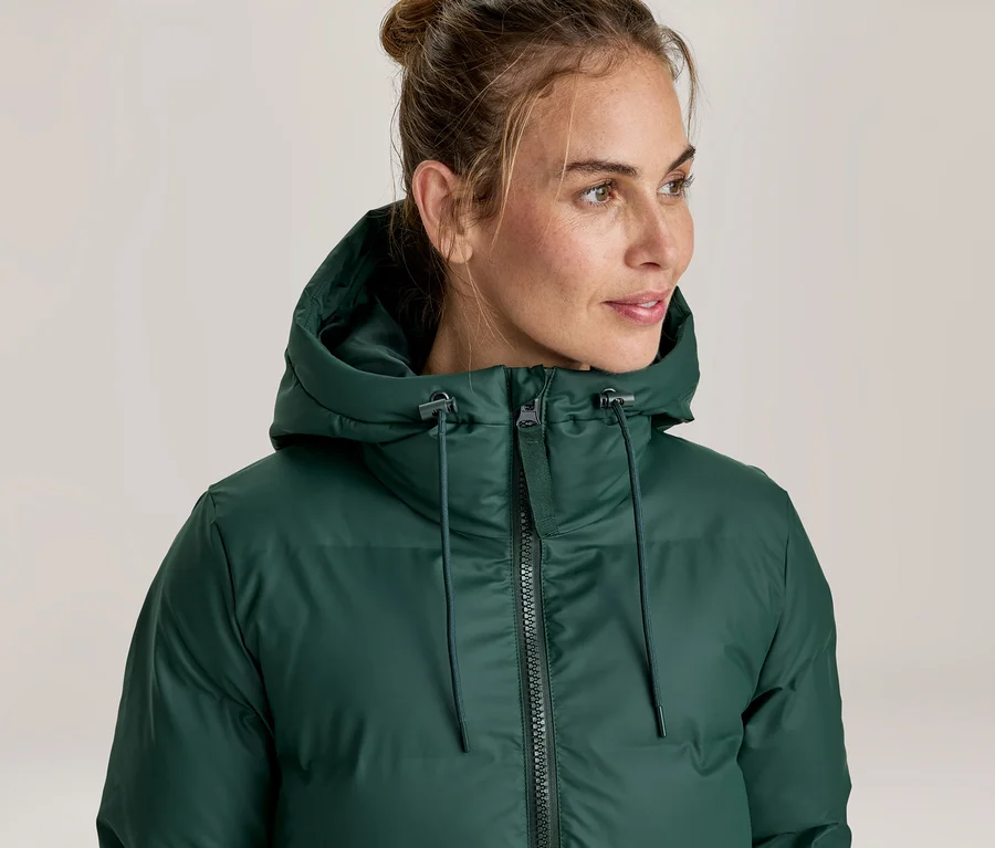Frau trägt eine dunkelgrüne Steppjacke mit Kapuze, blickt nach rechts. Ihr Haar ist zu einem Dutt gebunden.