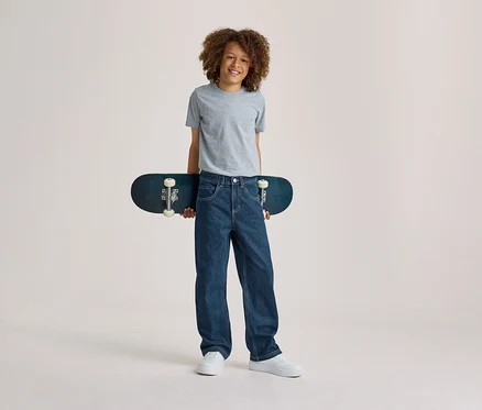 Lächelnder Junge mit lockigem Haar, der ein graues T-Shirt, blaue Jeans und weiße Sneaker trägt und ein dunkelblaues Skateboard hinter seinem Rücken hält.