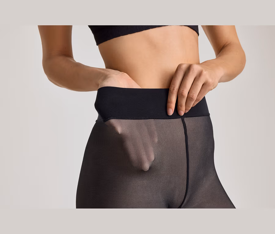 Nahaufnahme einer Person in transparenter schwarzer Strumpfhose und schwarzem Oberteil, eine Hand im Bund, durch den Stoff sichtbar, die andere Hand darauf.